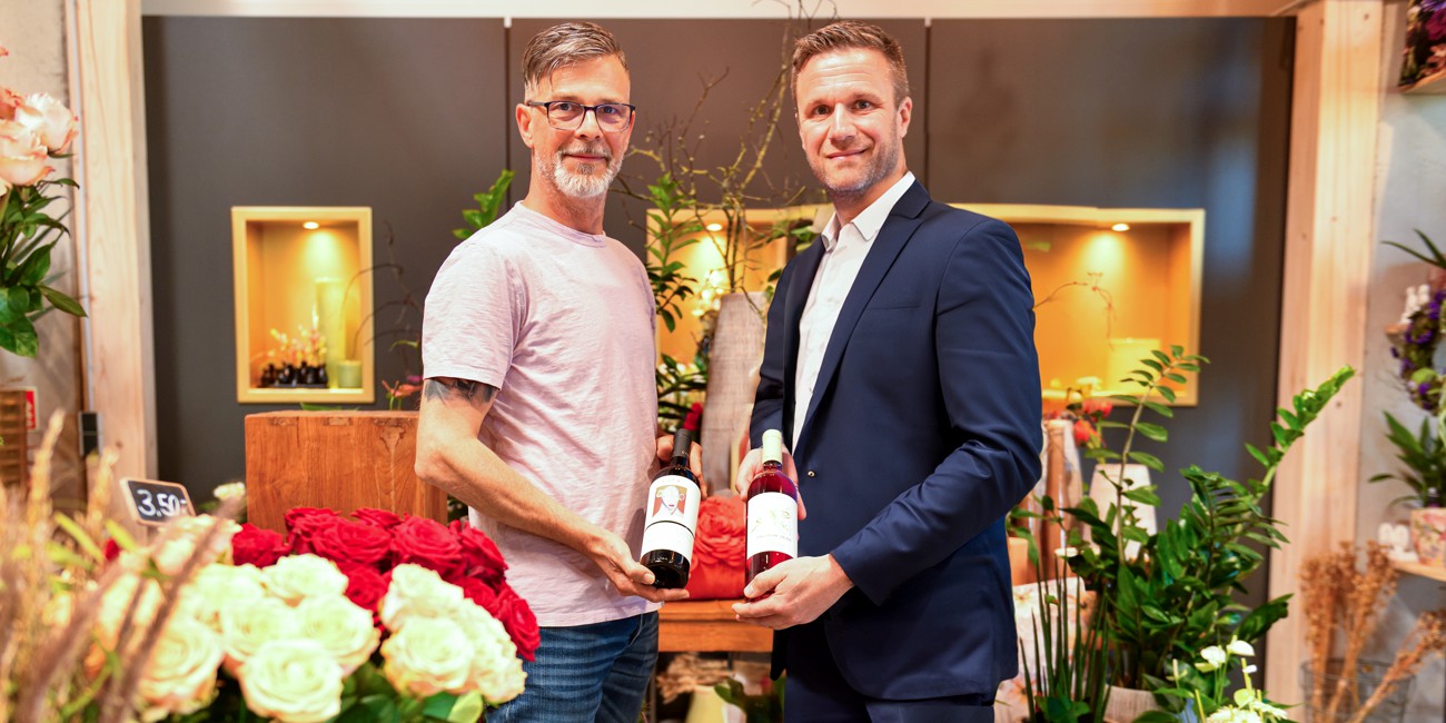 Zwei Männer stehen in einem Geschäft für Floristik und Feinkost, einer im Anzug, der andere in einem T-Shirt. Sie halten gemeinsam einen Geschenkkorb mit zwei Weinflaschen. Im Hintergrund sind Blumen und Pflanzen zu sehen.
