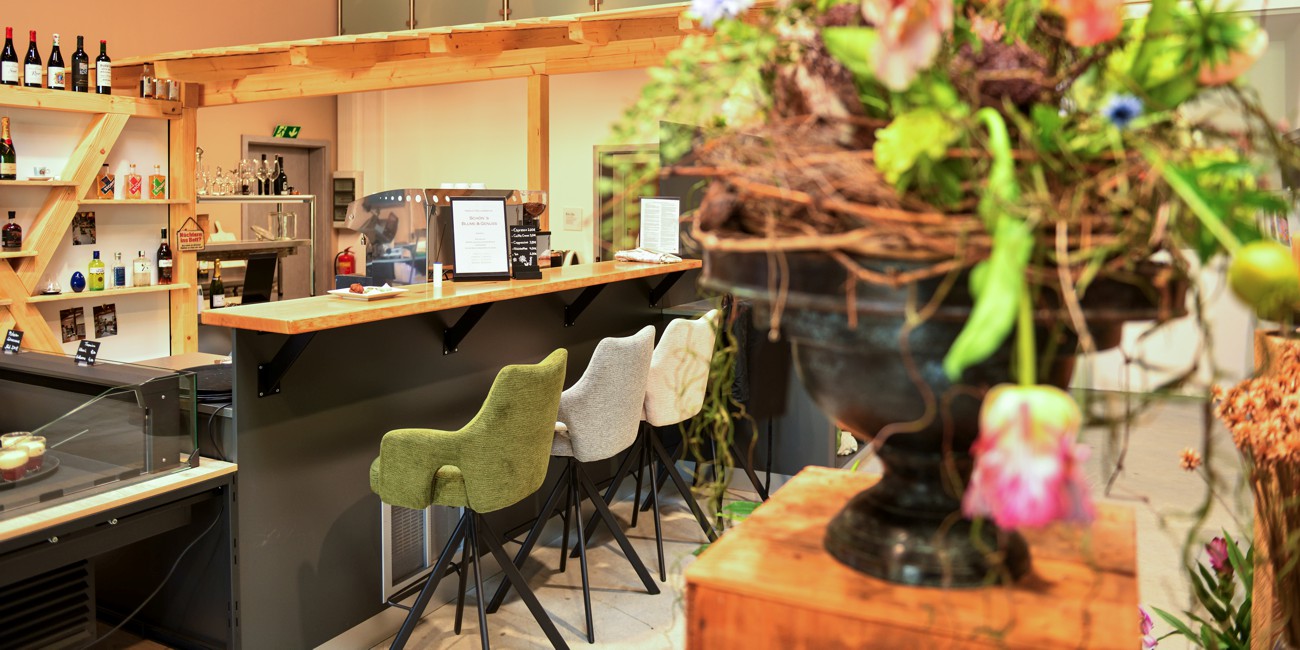 Gemütlicher Barbereich mit Theke aus Holz, verschiedenen Barhockern und Weinflaschen im Regal, im Vordergrund unscharf ein Blumengesteck.