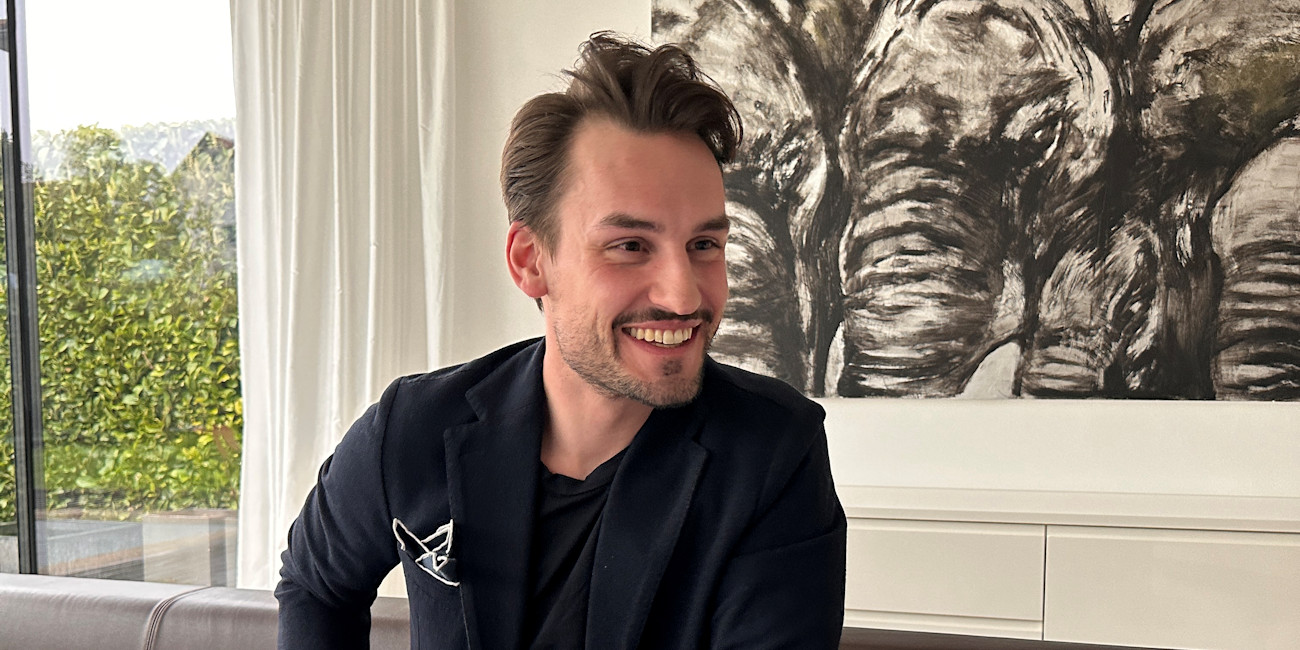 Alexander Noworzyn sitzt im Wohnzimmer und lächelt in die Kamera.