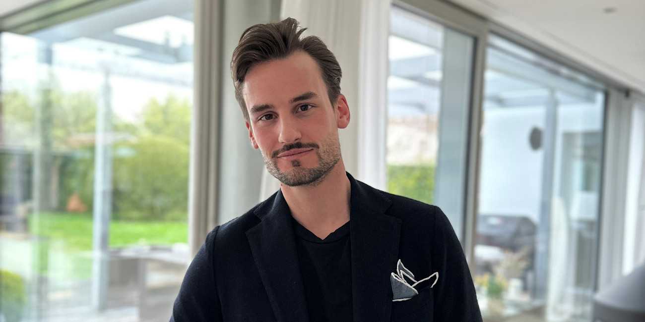 Alexander Noworzyn im schwarzen Jackett steht vor einer Fensterfront.