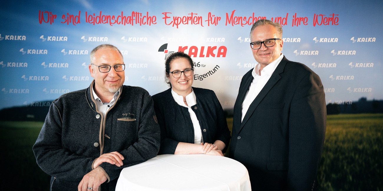Christian Flemisch, Eva Heißwolf und Jürgen Flemisch stehen vor einer Plakatwand mit der Aufschrift Wir sind leidenschaftliche Experten für Menscnen und ihre Werte - Kalka