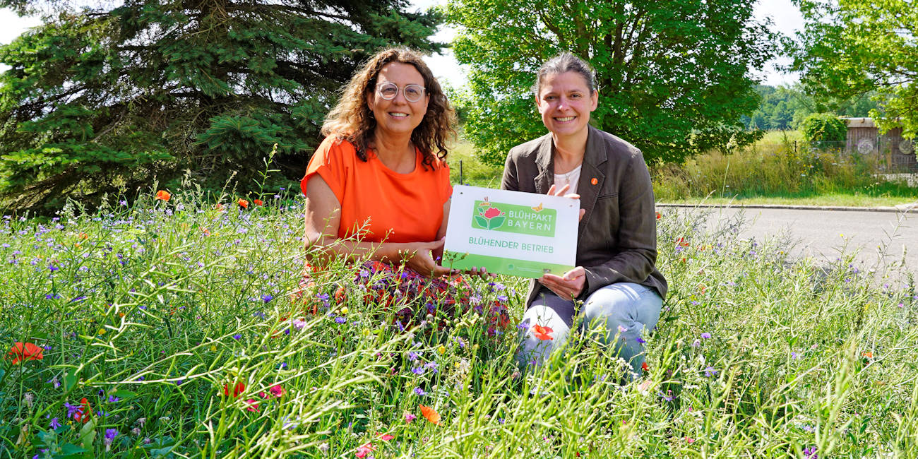 Miriam Albrecht und eine weitere Frau knien inmitten blühender Blumen mit einem Schild in der Hand, Blühpakt Bayern – blühender Betrieb.