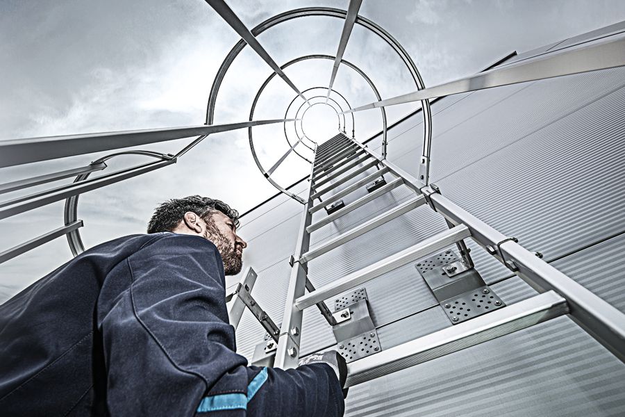 Ein Techniker steigt auf eine hoch ausgezogene Aluminiumleiter in eine industrielle Anlage – Blick nach oben durch die Konstruktion.
