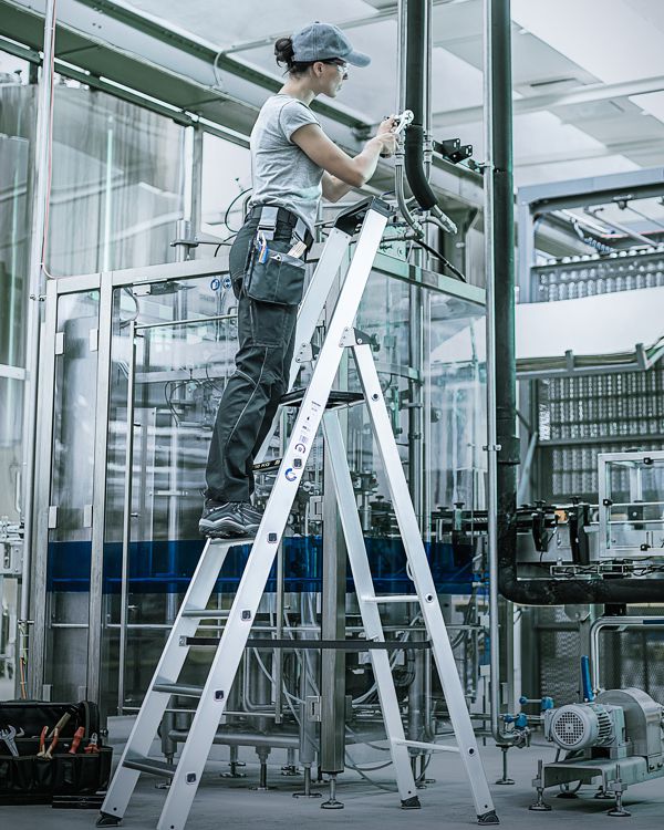 Eine Frau montiert Glaswände in einer Fertigungshalle – sie arbeitet auf einer Aluleiter mit Sicherheitsbügeln.