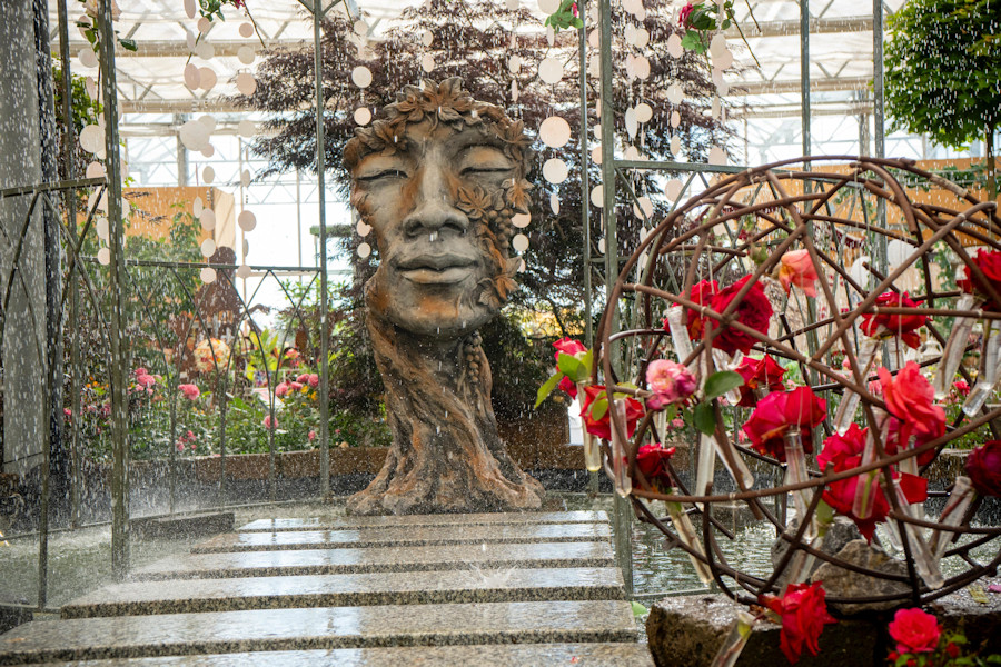 Ein Springbrunnen mit einer Blumenkugel und einem Gesicht mit geschlossenen Augen, das in einem Baumstamm eingelassen ist.