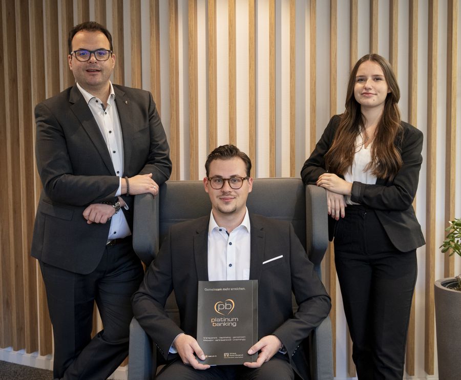 Private Banking Berater Christian Walburger und Maria Schmid, Assistenz PlatinumTeam stehen seitlich an einem Sessel, in dem Versicherungsexperte Ben Rieblinger sitzt mit einer Mappe von PlatinumBanking in der Hand.